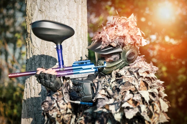 an airsoft sniper hiding behind tree aiming colorful gun wearing leafy ghillie outfit