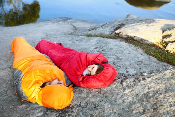 man in yellow sleeping bag and woman in red sleeping bag resting on cliff near lake