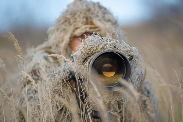 person in tan ghillie looking through large lens camera