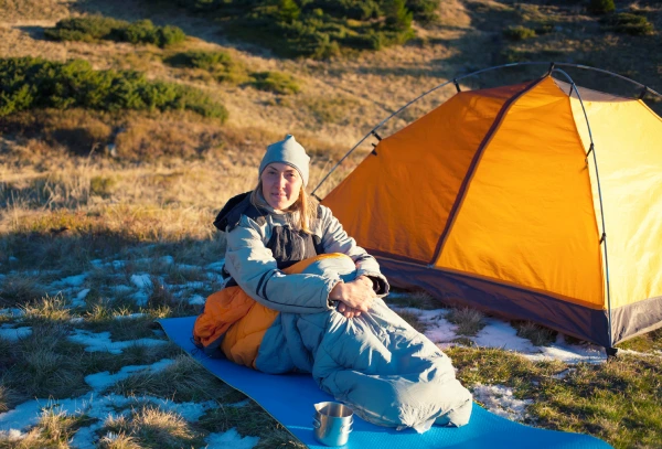 woman sitting in a sleeping bag on patchy snow ground next to orange tent