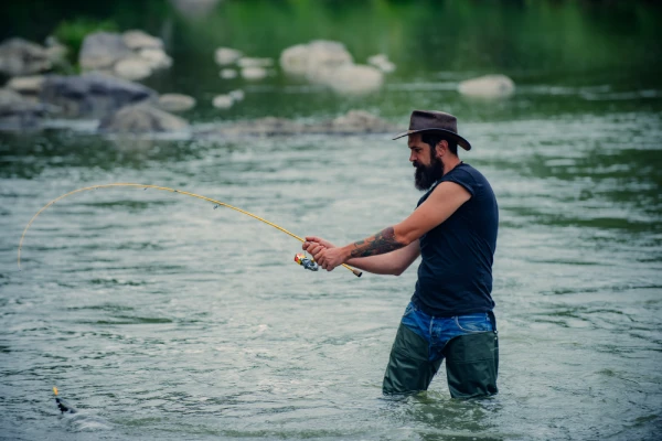 rugged bearded man in cowboy hat fishing with short yellow colored rod in river