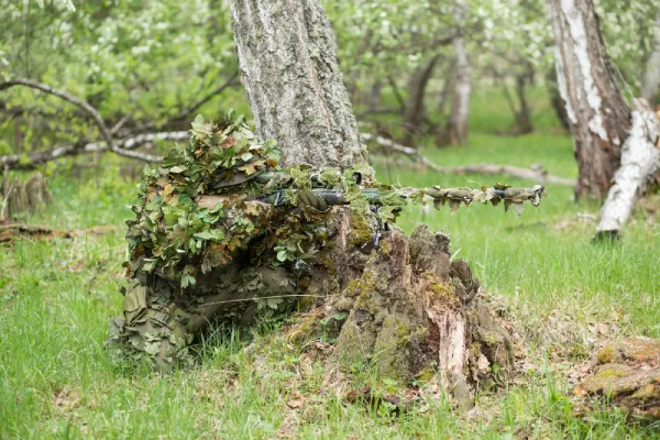 sniper wearing leafy ghillie crouching against tree pointing gun