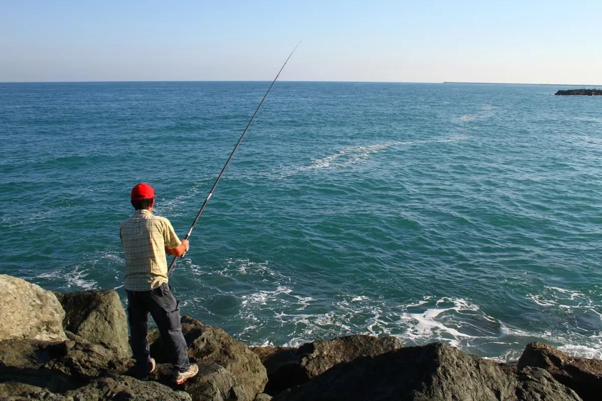 fisherman wearing red hat on rocks casting into ocean